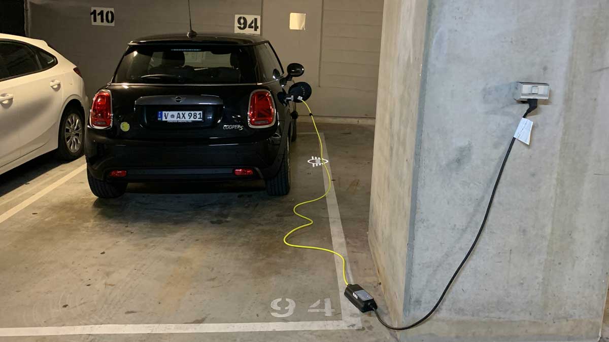 Electric car parked in an underground car park and plugged into a wall outlet with a charging cable running across the floor.