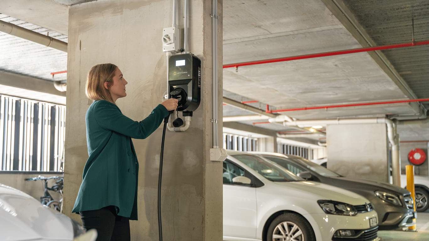 Person plugging an electric vehicle charging cable into a wall-mounted charger in an underground apartment car park, with parked cars in the background.