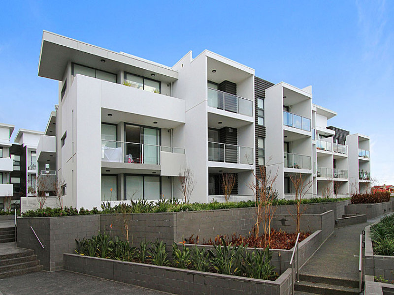 Modern three-storey apartment building with white exterior walls, glass balcony railings, and landscaped garden beds along a paved walkway.