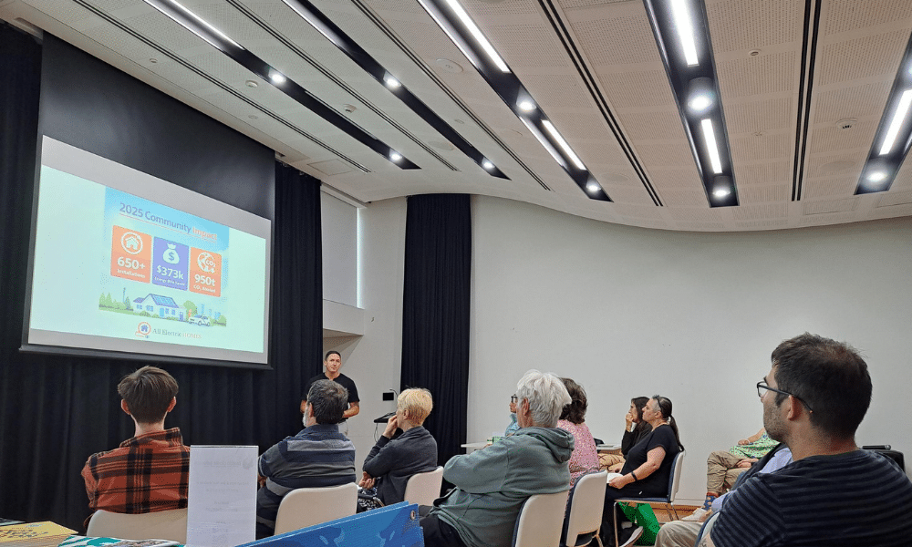 Presenter speaking to a seated audience at a community electrification event, with a presentation slide showing All Electric Homes’ 2025 community impact.