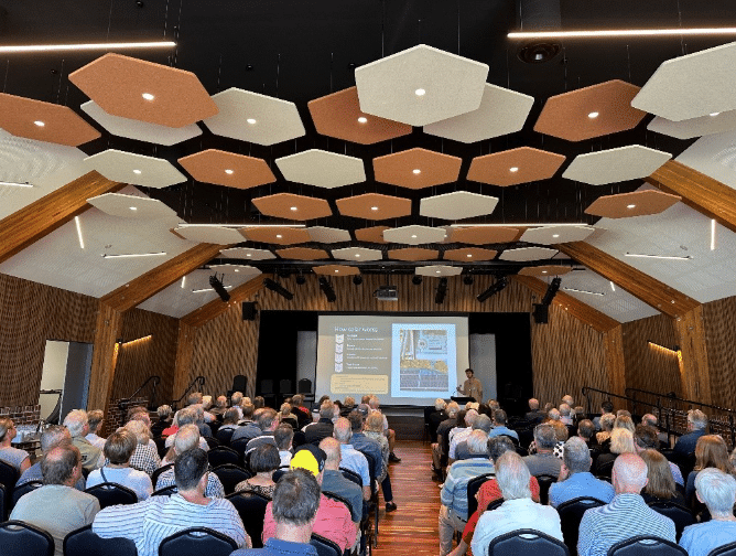 A large audience seated in a community hall watching a presentation on home energy upgrades, with a presenter speaking beside a projected slide.