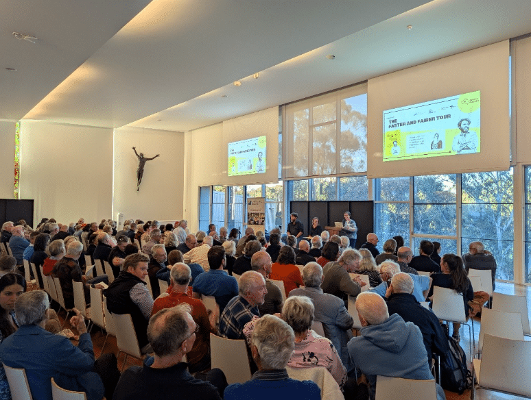 A large audience seated in a bright community venue watching a presentation titled “The Faster and Fairer Tour”, with speakers at the front of the room.